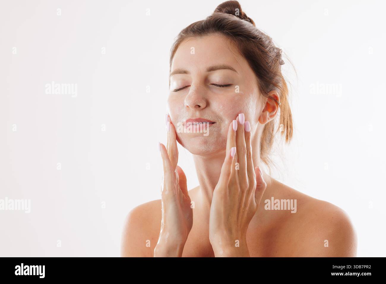 Jeune femme à la peau éclatante se lavant le visage avec un nettoyant doux, profitant d'une routine de soins de la peau, isolée sur un fond blanc. Banque D'Images