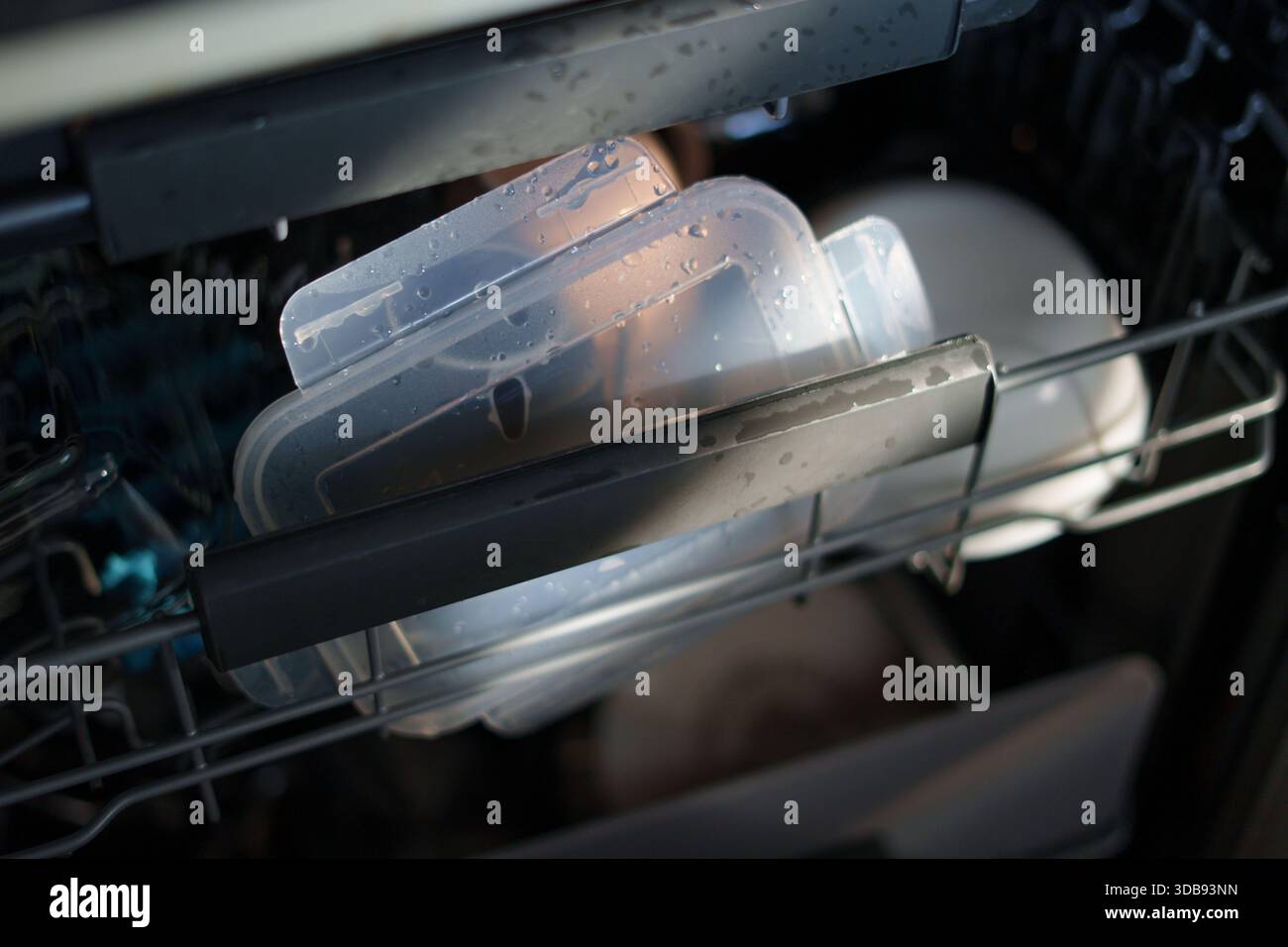 vue rapprochée d'un panier de lave-vaisselle chargé de récipients et d'assiettes en plastique transparents humides, éclairés par une lumière naturelle douce, mettant en évidence les gouttelettes Banque D'Images