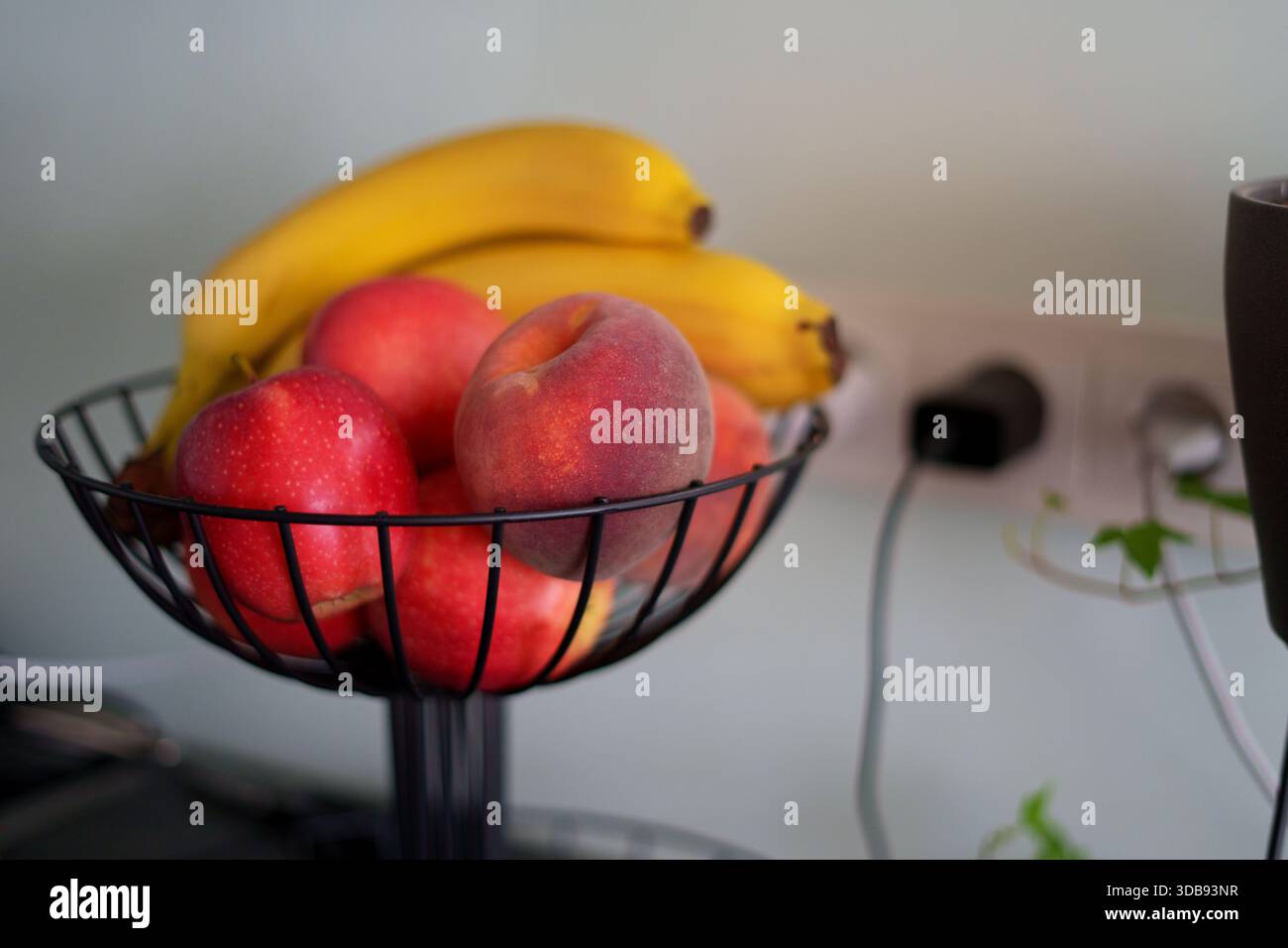un arrangement de fruits vibrant avec des pommes mûres, des bananes et une pêche dans un panier en fil de fer noir sur un fond de cuisine neutre, créant un a frais Banque D'Images