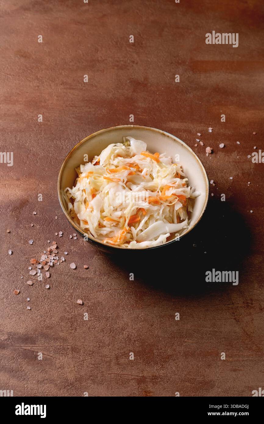 Chou choucroute traditionnel avec des carottes dans un bol en céramique. Sel de mer à proximité. Nourriture saine, produits fermentés. Fond texturé marron, espace de copie. Banque D'Images