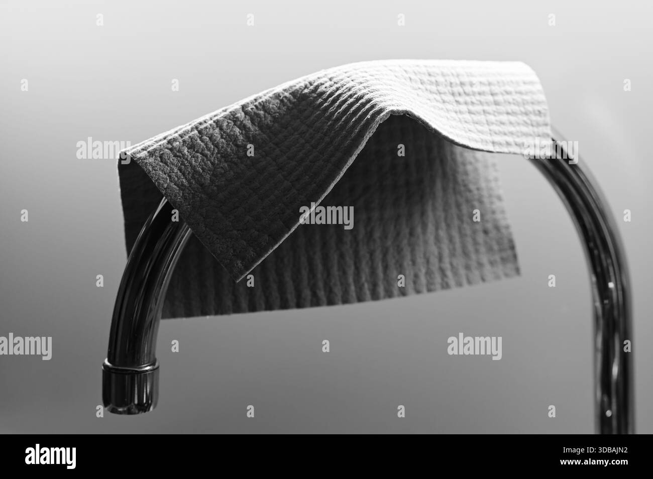 Une nature morte minimaliste en noir et blanc montrant un chiffon de nettoyage texturé drapé sur une barre chromée incurvée, soulignant les détails du tissu, les ombres douces, et un Banque D'Images