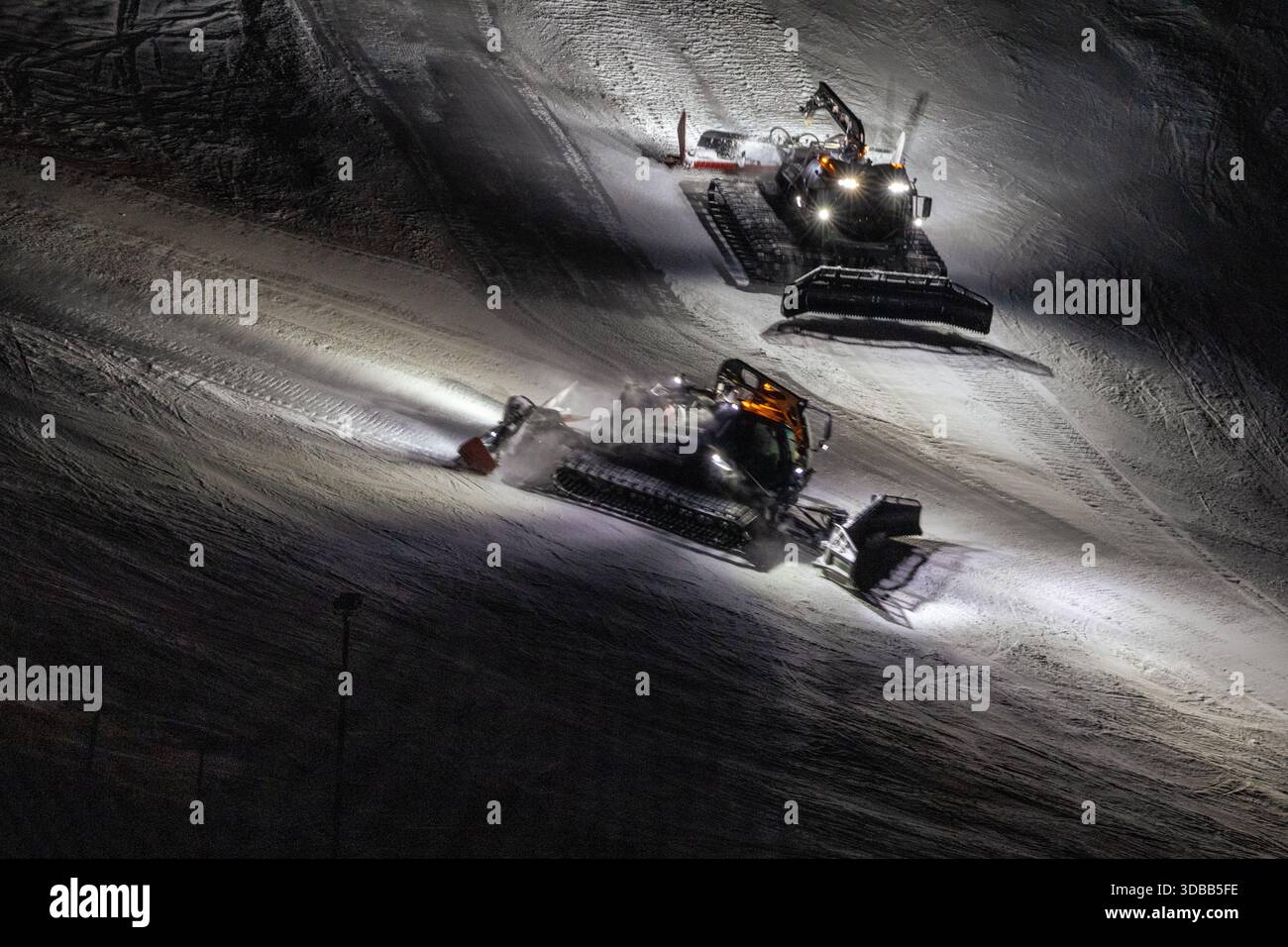 Deux tondeuses puissantes travaillent avec diligence sur une piste de ski sous le couvert de la nuit. Leurs lumières brillantes illuminent la neige fraîchement entretenue, créant ainsi Banque D'Images