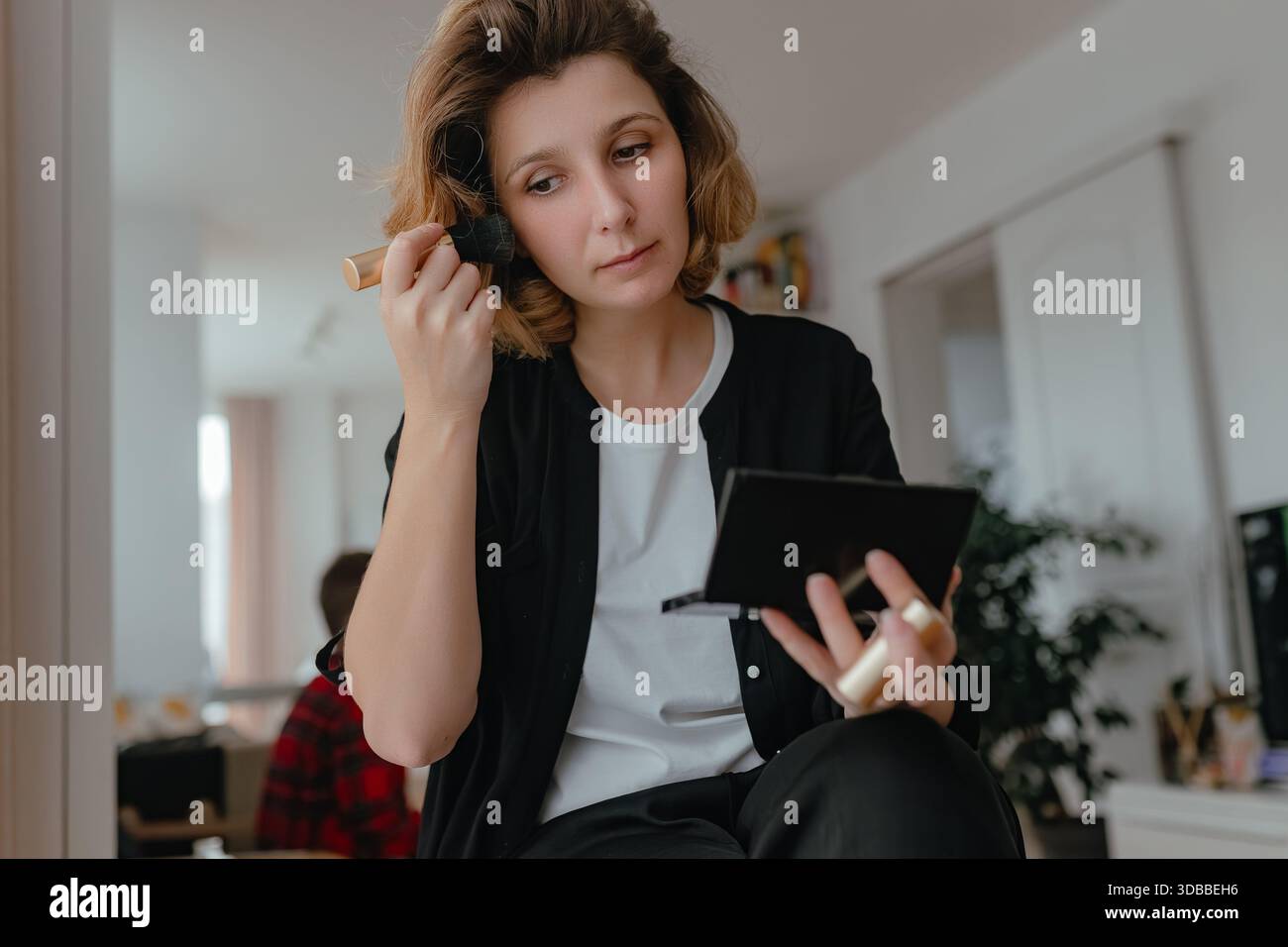 Femme appliquant le contour avec le pinceau de maquillage, lumière douce intérieure, routine de beauté naturelle, vêtements décontractés, expression concentrée, éléments intérieurs chauds. Banque D'Images