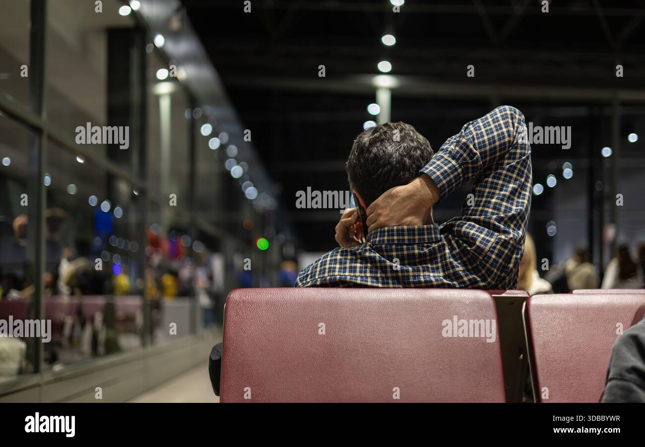 homme solitaire en chemise à carreaux attend un vol retardé dans un terminal d'aéroport sombre et moderne, parlant au téléphone avec la main derrière le cou. Banque D'Images