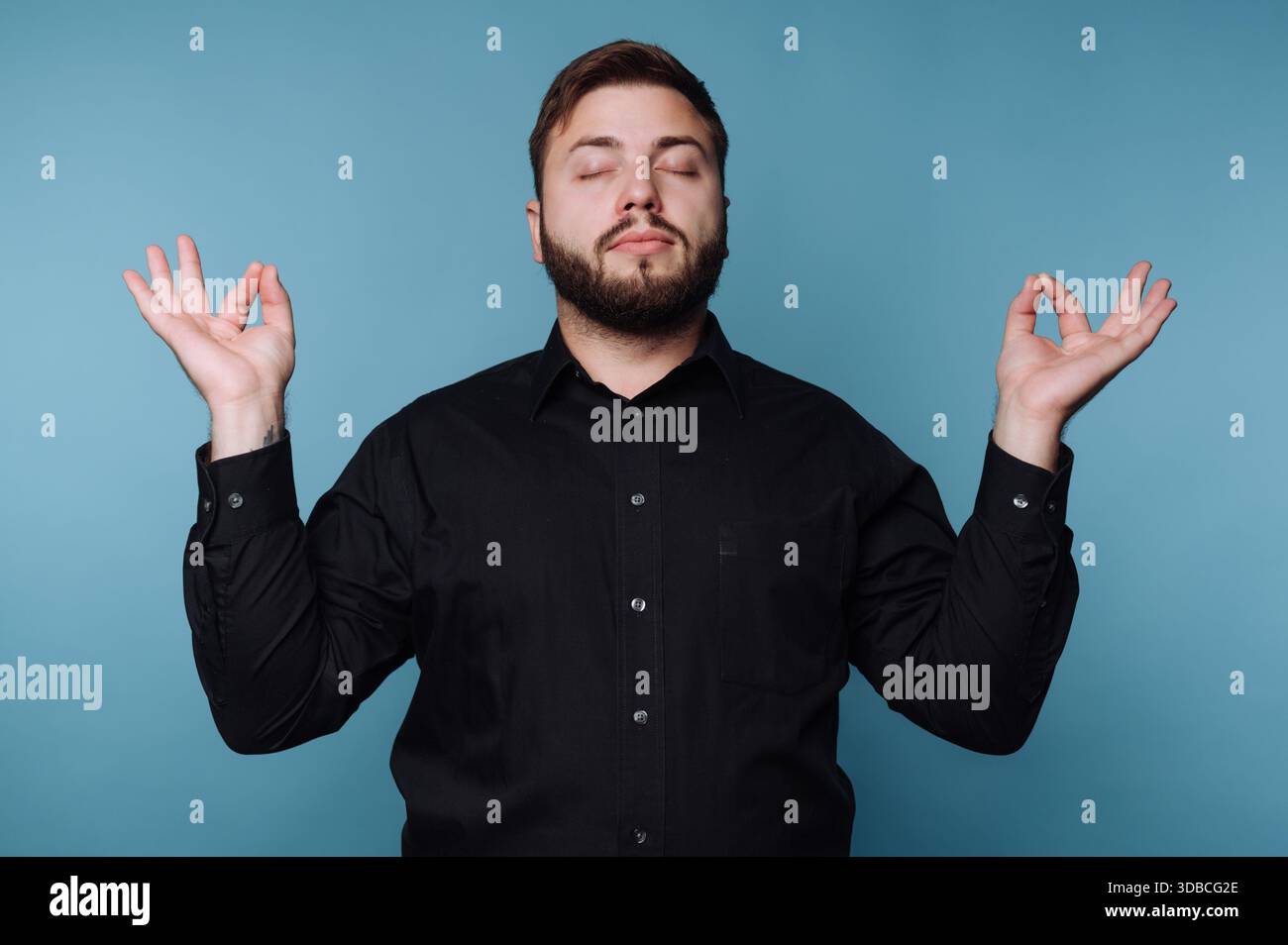 Un homme pratique la méditation les yeux fermés, les mains dans un geste de paix. Il est à l'intérieur contre un mur bleu. C'est l'après-midi et il semble focuse Banque D'Images