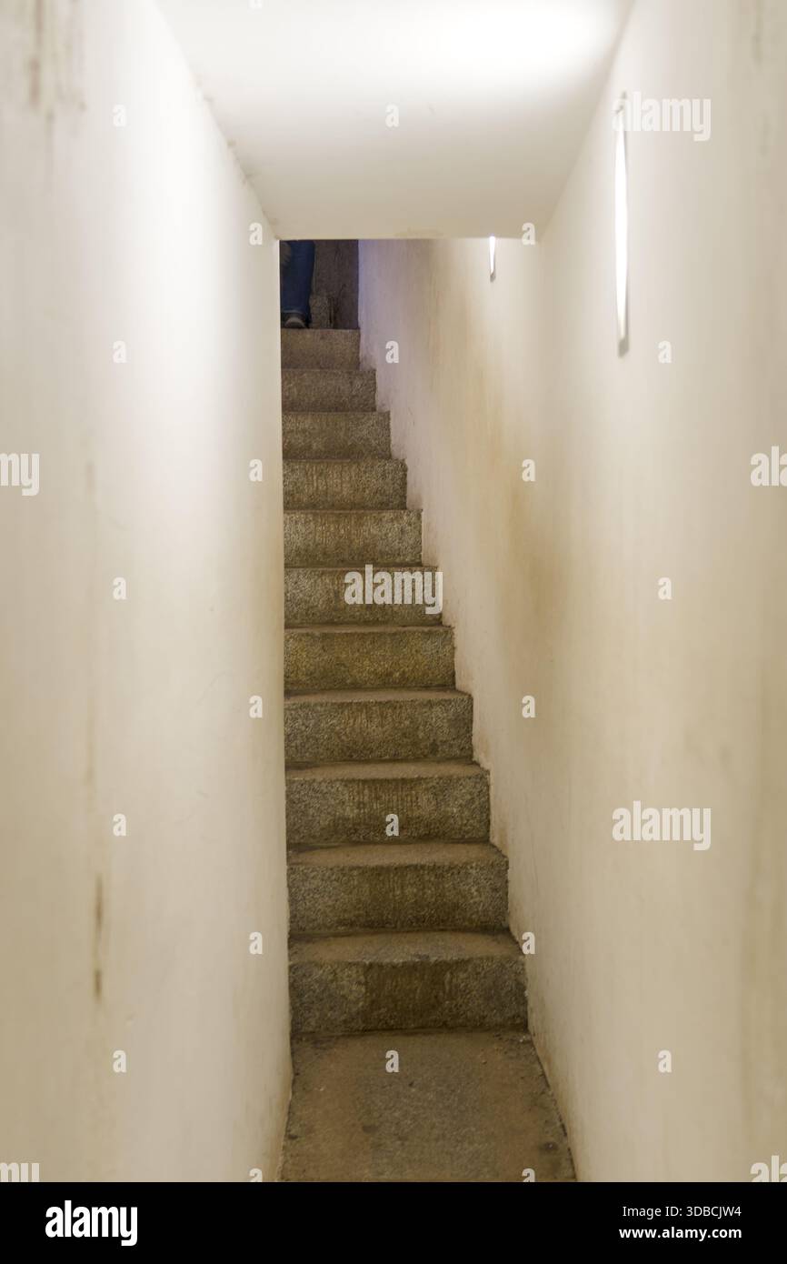 Un escalier en pierre serré mène vers le haut à travers un couloir étroit, mettant l'accent sur l'espace confiné et la texture architecturale brute Banque D'Images