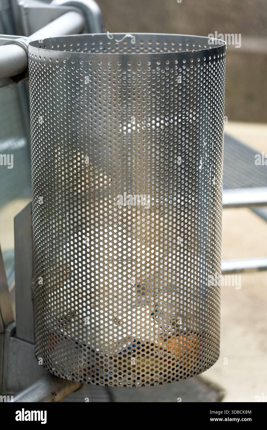 Poubelle en métal perforé debout dans un environnement urbain, montrant un design de rue fonctionnel avec un aspect industriel et minimaliste. Banque D'Images
