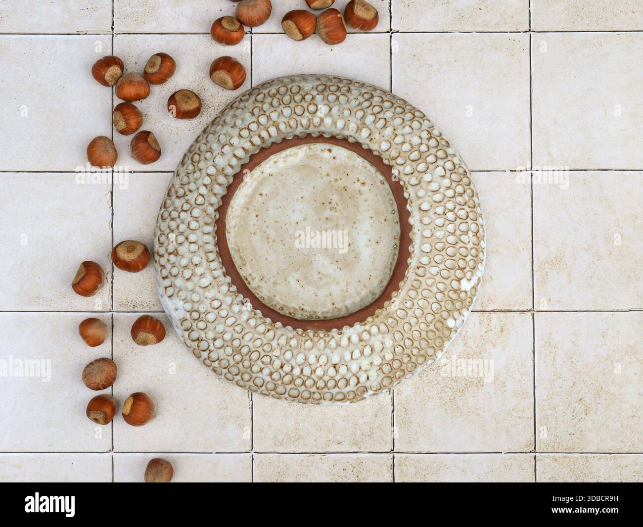Vue de dessus d'un bol en céramique fait à la main avec texture circulaire sculptée placé sur la surface carrelée. Composition de nature morte avec des noisettes disposées à proximité, photo Banque D'Images