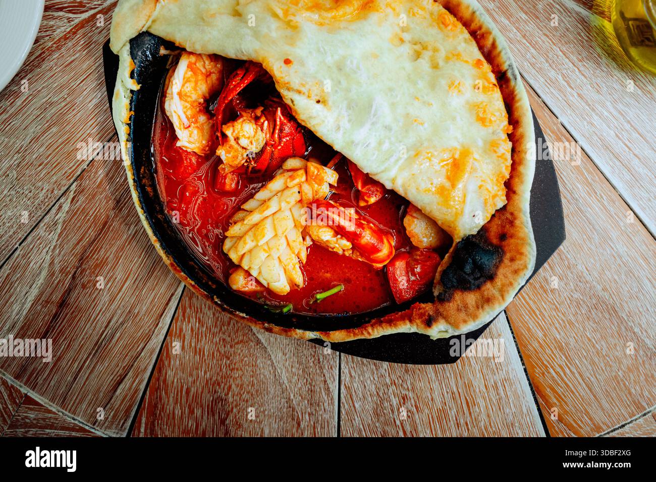 Le ragoût de fruits de mer en croute présente des crevettes, des calmars et du crabe dans une poêle en fonte recouverte de pain sur une table en bois à côté de l'huile. Annonce de poivrons rouges éclatants Banque D'Images