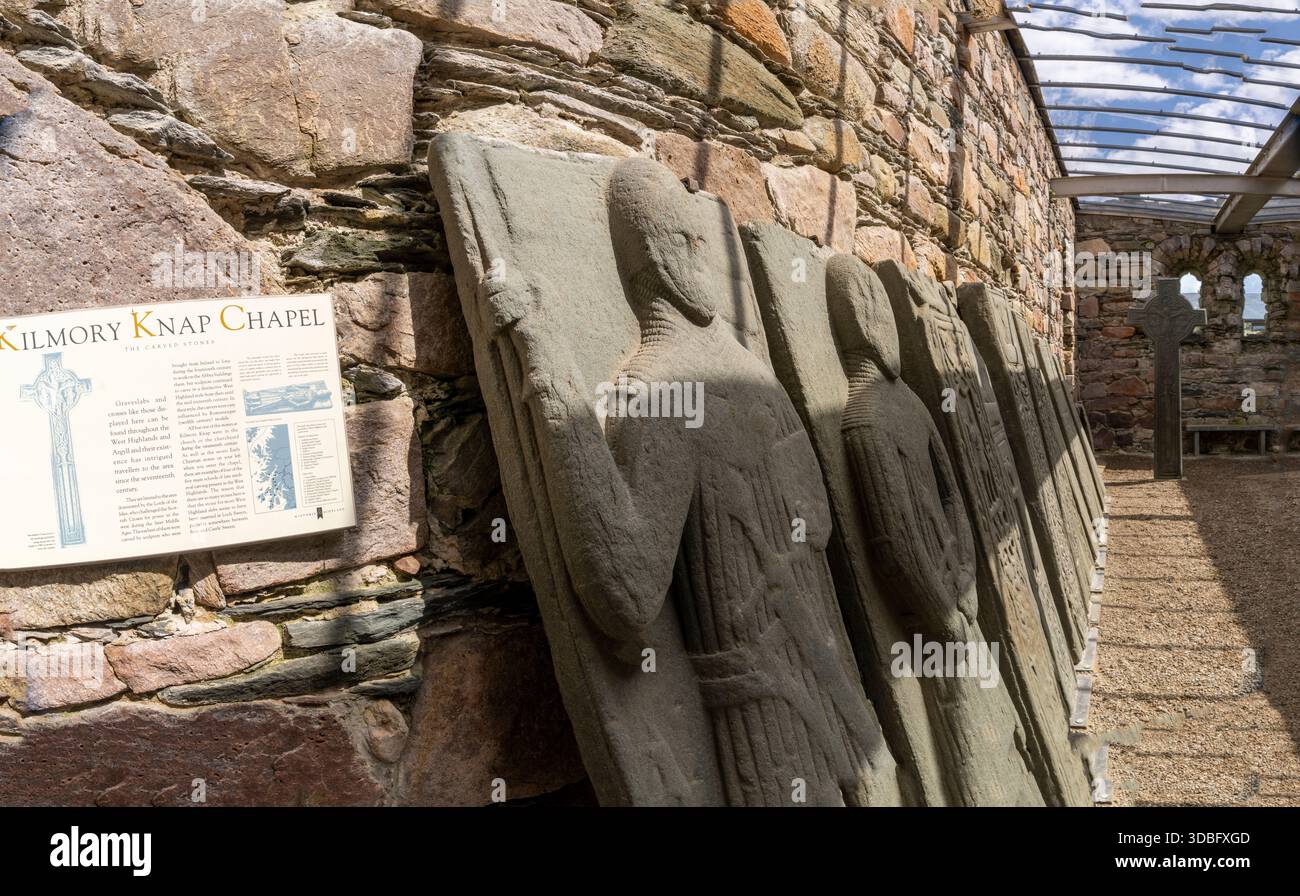 Une rangée de vieilles figures en pierre sculptée se dresse contre un mur de pierre accidenté à côté du site de la chapelle Kilmory Knap, avec une arche en bois et en pierre et des informations Banque D'Images