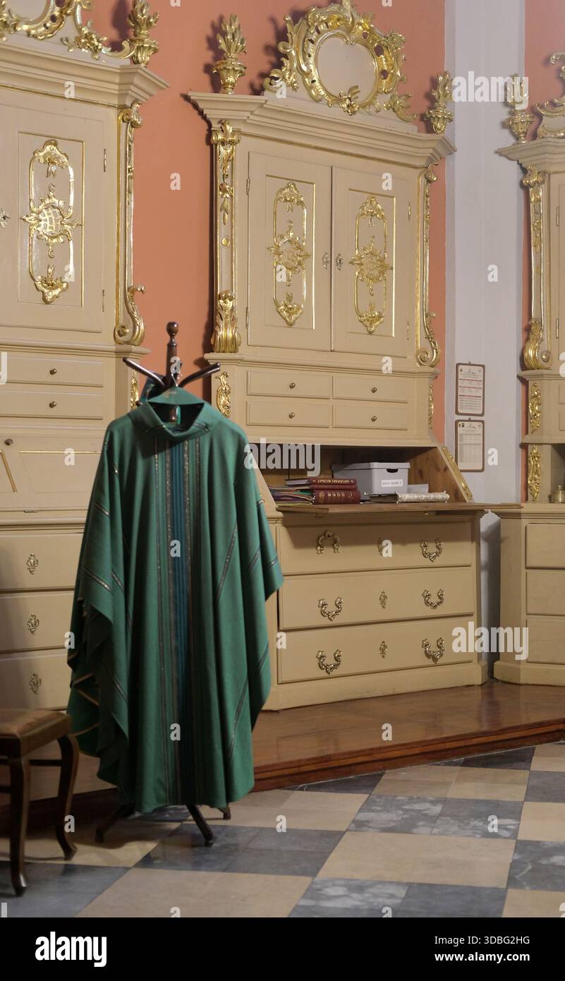 Vue intérieure d'un chasuble liturgique vert suspendu sur un support dans une sacristie avec des armoires ornées de couleur crème, des moulures décoratives dorées, des murs roses Banque D'Images