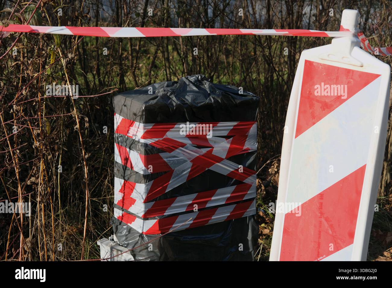 Scène extérieure montrant un objet rectangulaire enveloppé dans du plastique noir et du ruban barrière rouge-blanc, panneau d'avertissement à côté, végétation sèche, lumière du jour Banque D'Images