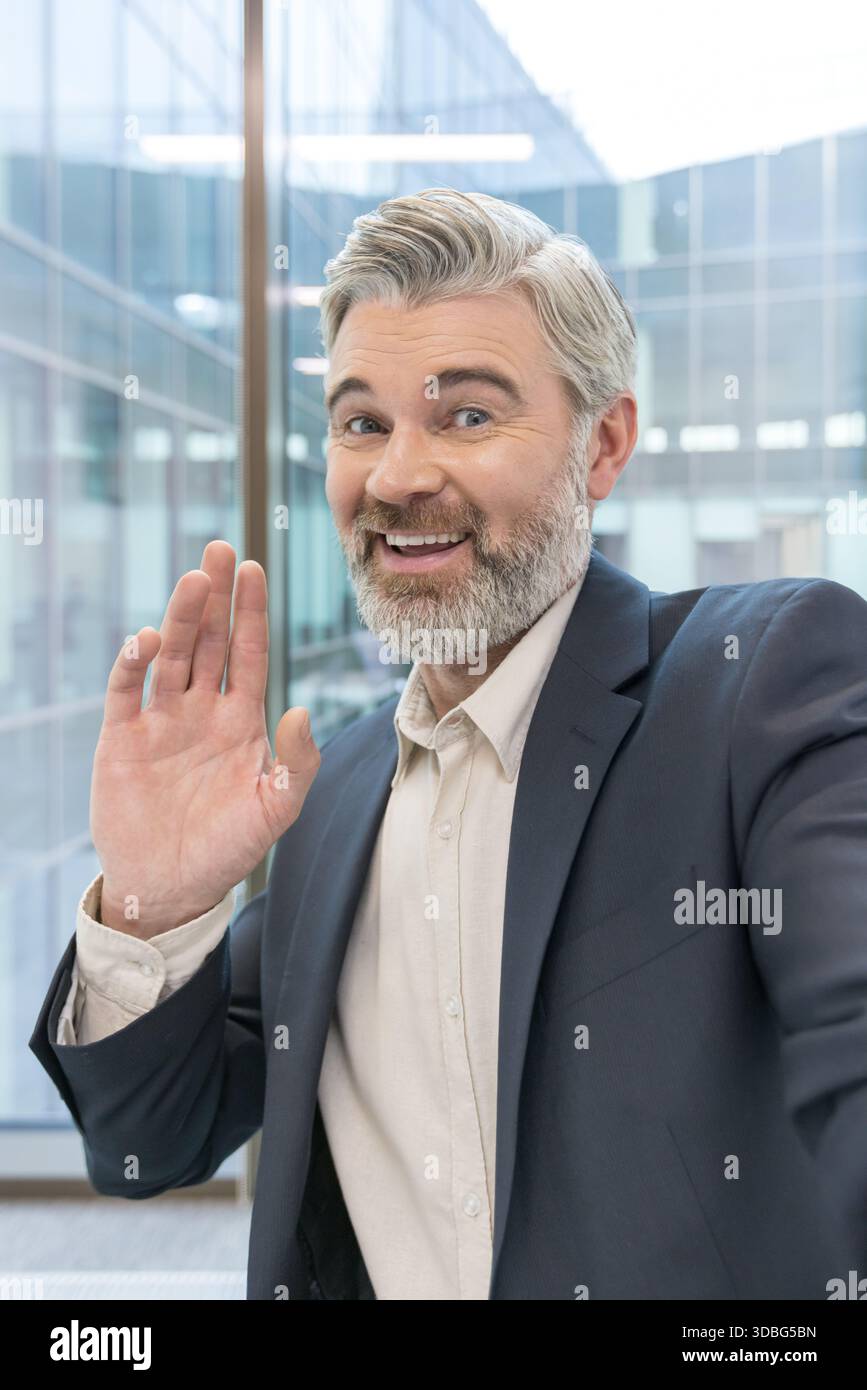 Homme d'affaires senior avec une barbe grise et une tenue professionnelle souriant joyeusement tout en agitant une main amicale à la caméra, debout dans un environnement de bureau moderne Banque D'Images