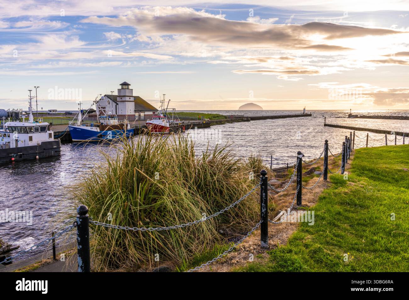 Scène pittoresque du port de Girvan avec des bateaux amarrés le long du quai sous un ciel de coucher de soleil et une côte herbeuse. Banque D'Images