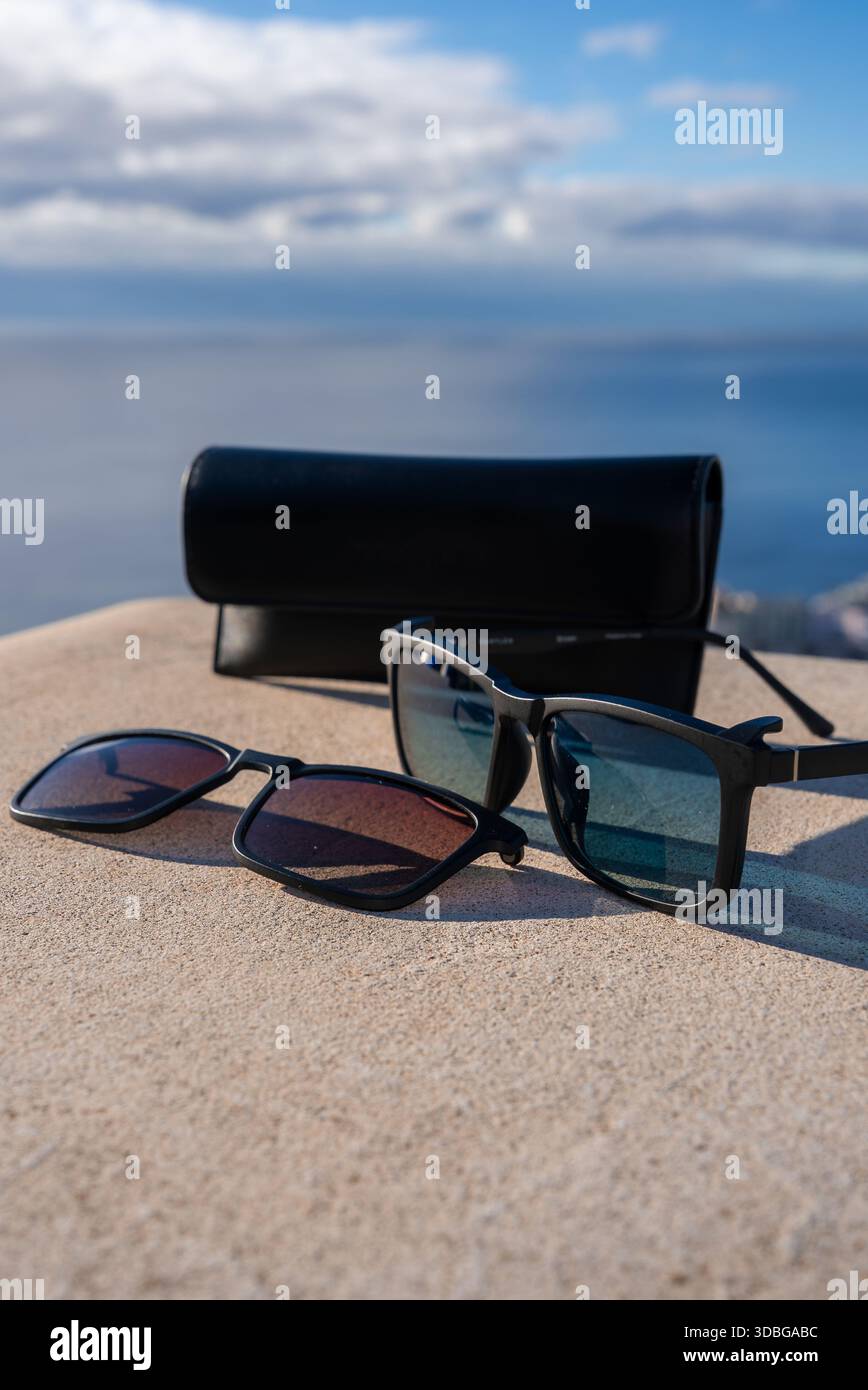 Deux paires de lunettes de soleil encadrées noires reposent sur un rebord de pierre ensoleillé avec un boîtier noir, près de Funchal à Madère, Portugal, sous un ciel partiellement nuageux par t Banque D'Images