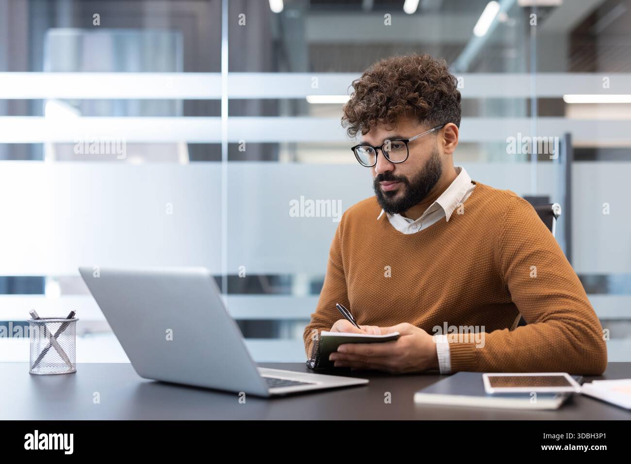 Jeune homme aux cheveux bouclés et à la barbe portant des lunettes, concentré sur l'apprentissage en ligne à partir de son ordinateur portable, prenant des notes dans un ordinateur portable à un bureau dans un espace de travail moderne Banque D'Images