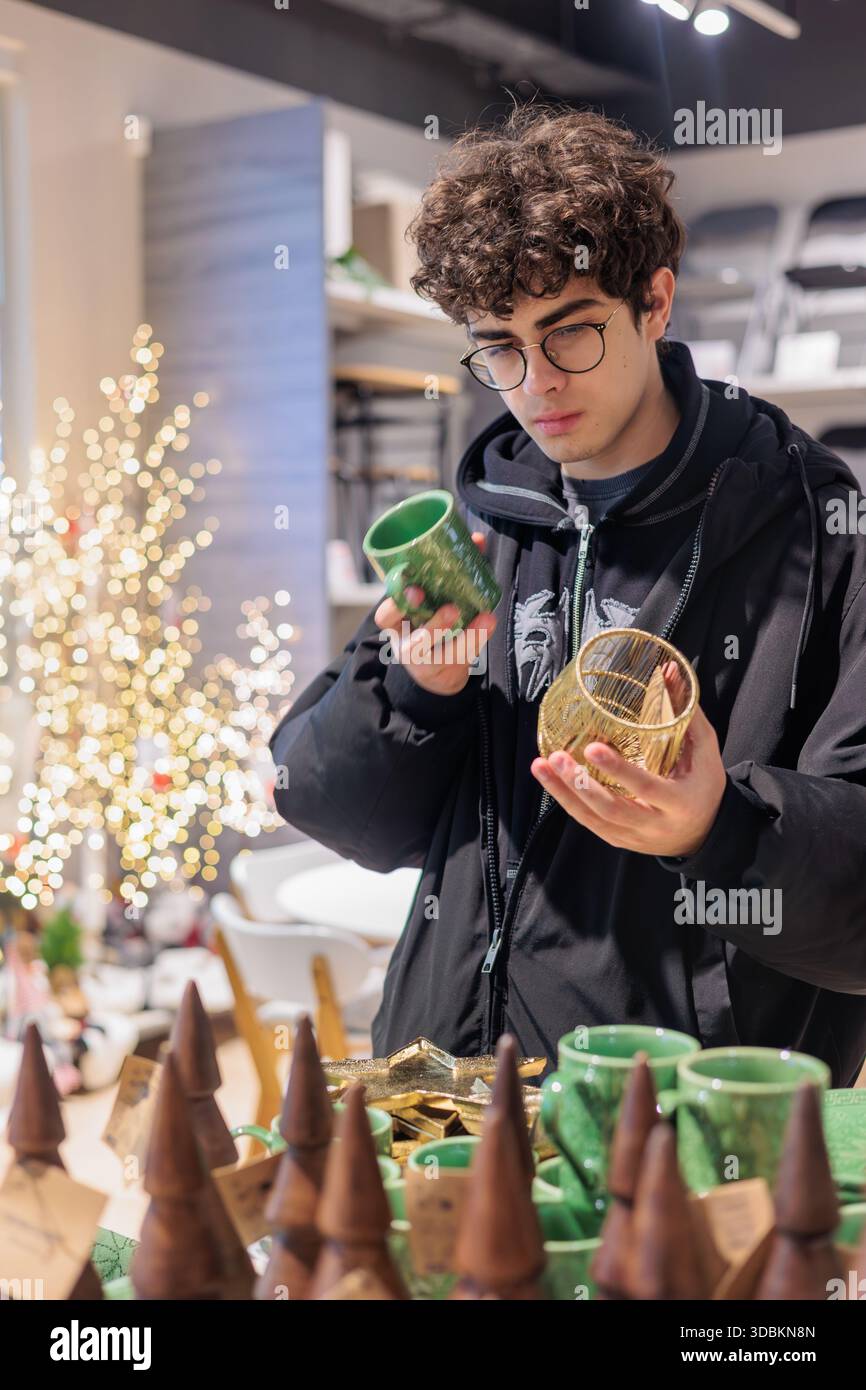 Adolescent choisissant une tasse de Noël et un décor en verre festif pendant les achats de vacances. Jeune homme comparant la tasse des fêtes et l'article en verre décoratif à Christma Banque D'Images