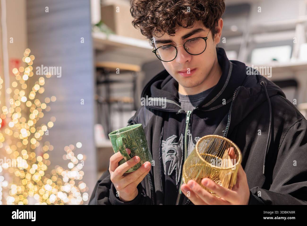 Adolescent choisissant une tasse de Noël et un décor en verre festif pendant les achats de vacances. Jeune homme comparant la tasse des fêtes et l'article en verre décoratif à Christma Banque D'Images