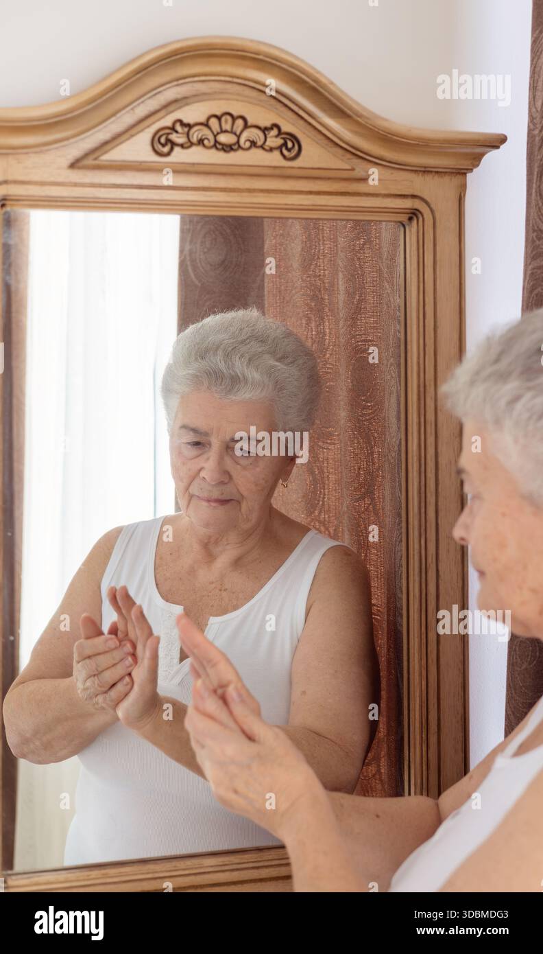 Femme âgée appliquant la crème pour les mains et massant ses mains devant le miroir – soins de la peau, soins personnels et routine beauté pour la peau mature. Banque D'Images
