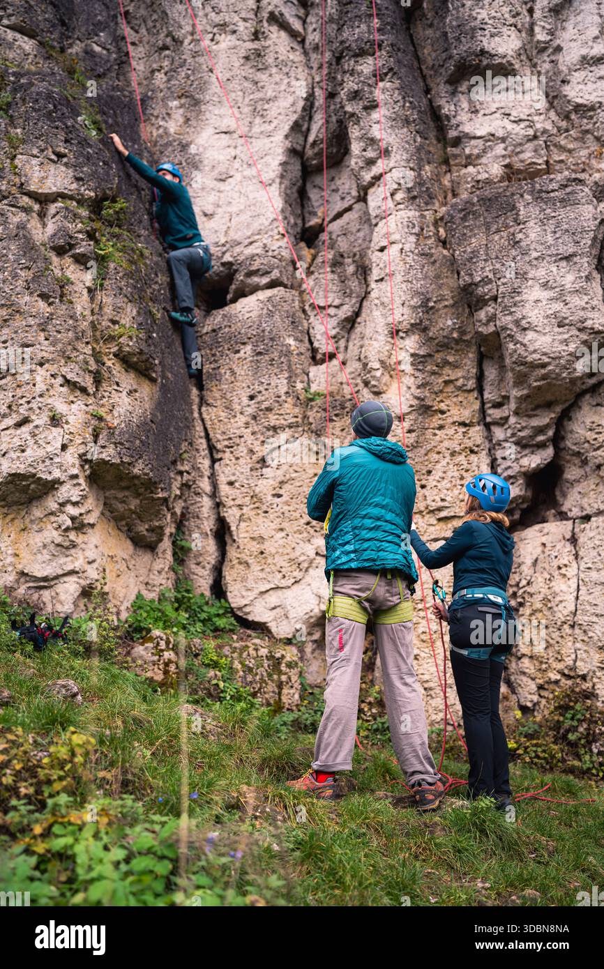 Un grimpeur est sécurisé par son partenaire de corde tout en grimpant sur le rocher. Un autre grimpeur avec un harnais d'escalade regarde le grimpeur. Banque D'Images