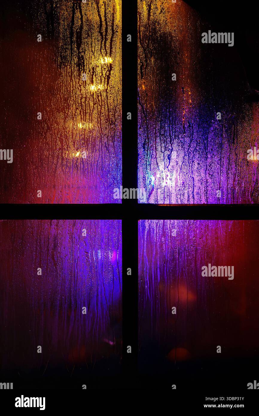 Une vitre sombre avec des gouttelettes de pluie la nuit. Lumières de noël jaunes et bleues floues à travers le verre humide recouvert de gouttes de condensat. Banque D'Images