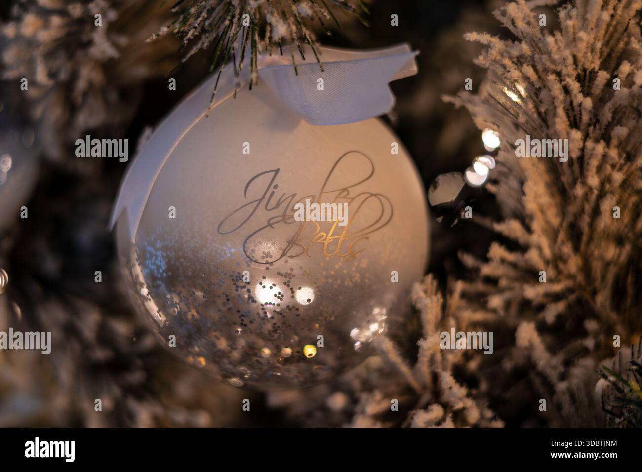 Close-up d'un ornement de Noël en verre blanc et clair avec des paillettes argentées et le texte "Jingle Bells", suspendu à un sapin artificiel givré. Banque D'Images