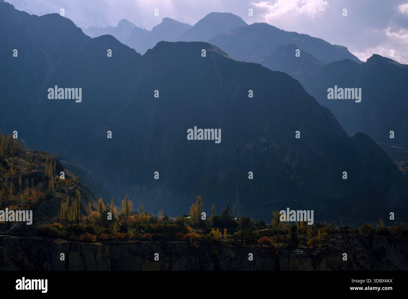 Vue sur les montagnes imposantes et brumeuses projetant de longues ombres sur la vallée, avec des arbres clairsemés accrochés aux pentes de Hunza, Gilgit Baltistan, Pakistan. Banque D'Images