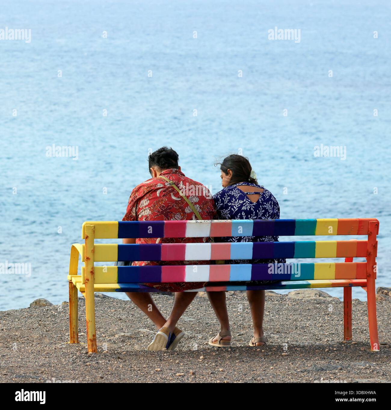Couple coloré assis sur un banc couleur arc-en-ciel regardant vers la mer, Fuerteventura, îles Canaries, Espagne. Prise en novembre 2025 Banque D'Images