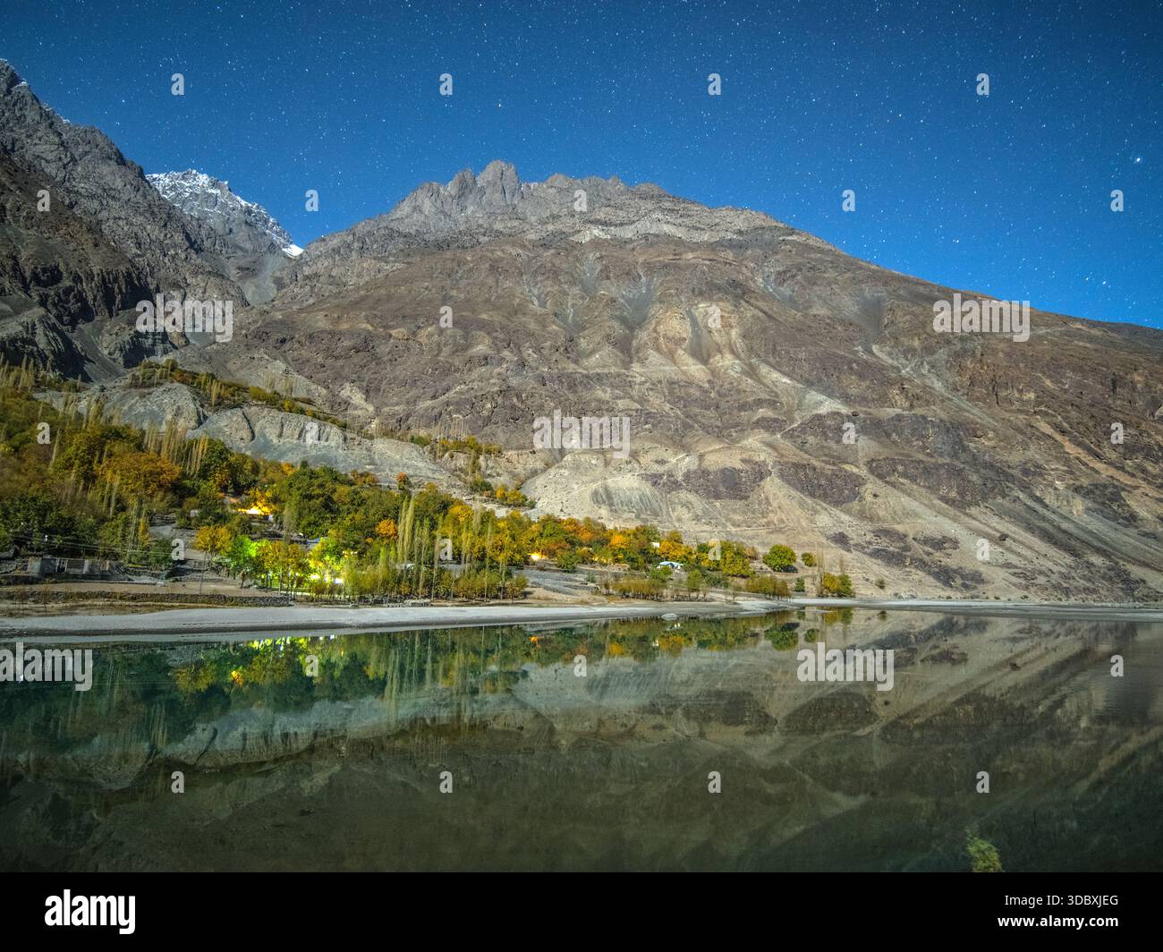 Vue sur un lac serein reflétant le ciel étoilé et les montagnes imposantes, niché à côté d'un village éclairé par des lumières chaudes, Gupis, Gilgit Baltistan, Banque D'Images