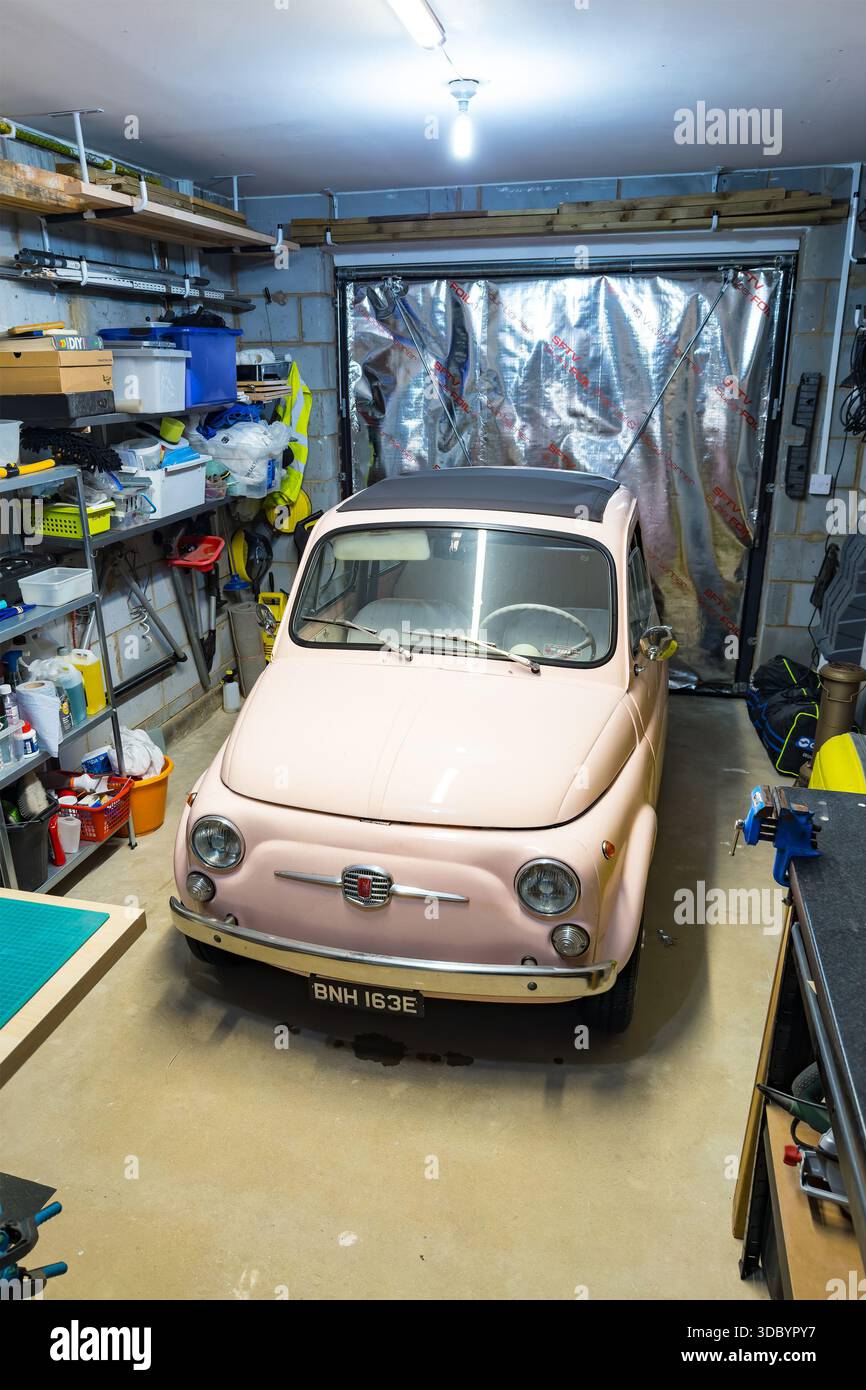 Vintage rose Fiat 500 garé à l'intérieur d'un garage de maison encombré rempli d'outils, d'étagères et de matériel d'atelier sous éclairage électrique Banque D'Images