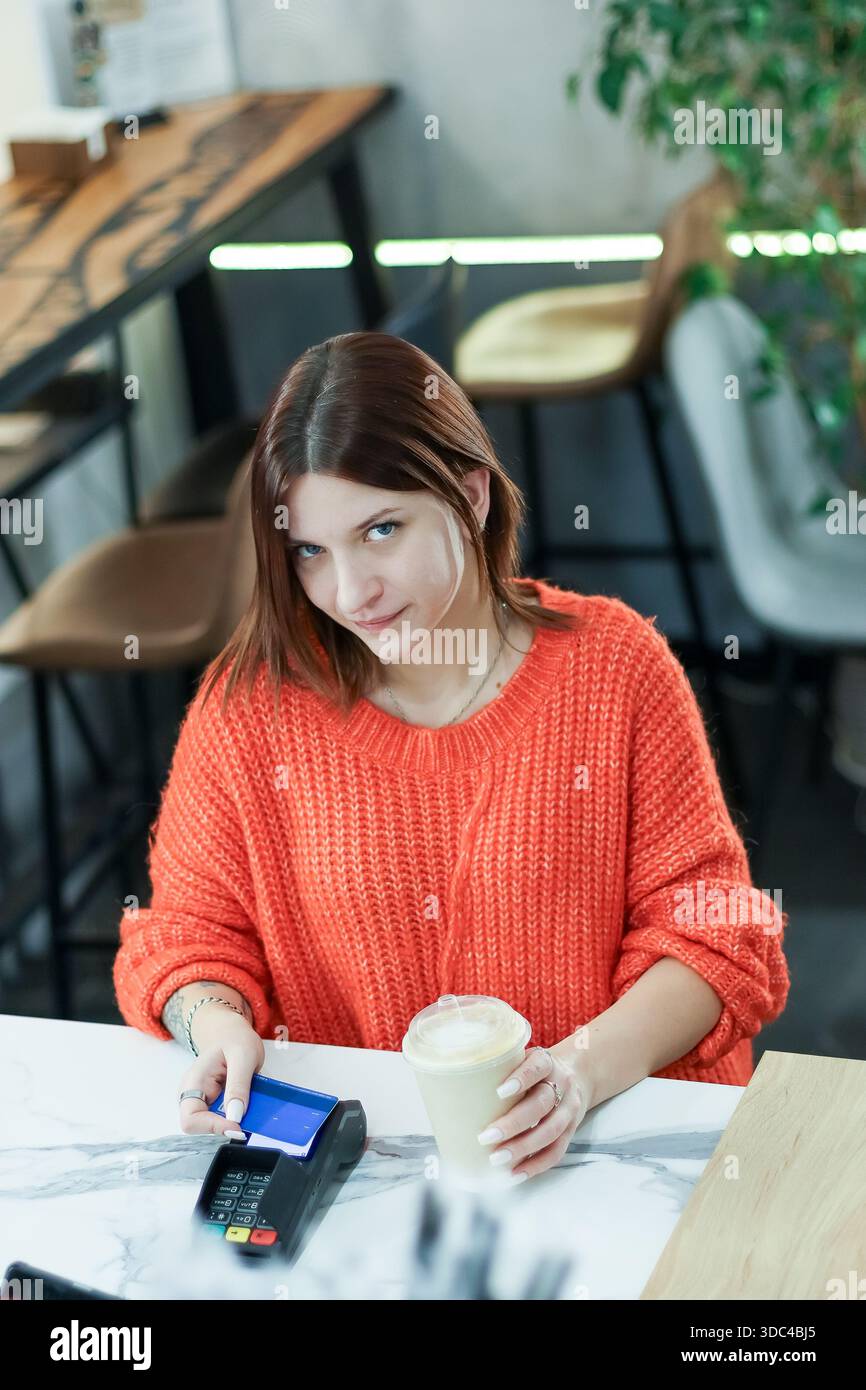 Une jeune femme souriante avec bob coupe de cheveux en tricot orange paie avec une carte bleue au comptoir du café, tenant son café à emporter. Banque D'Images