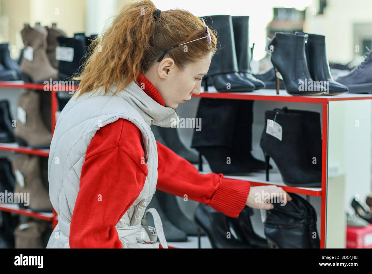 Vue latérale d'une cliente cherchant une bottine noire sur une étagère rouge dans un magasin de chaussures très fréquenté. Banque D'Images