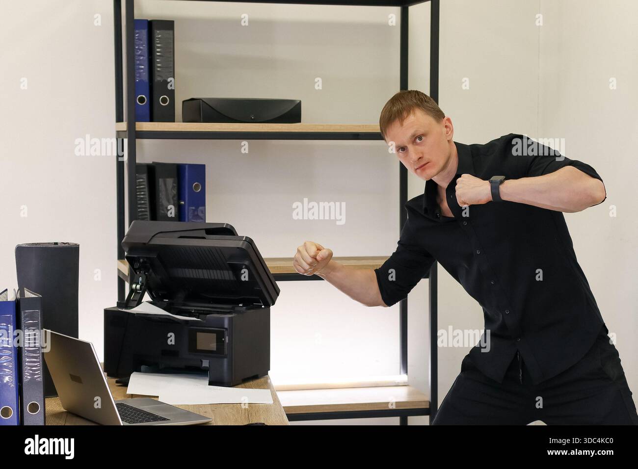 Homme frustré en chemise noire serrant le poing agressivement vers une imprimante défectueuse dans un espace de travail de bureau moderne. Banque D'Images