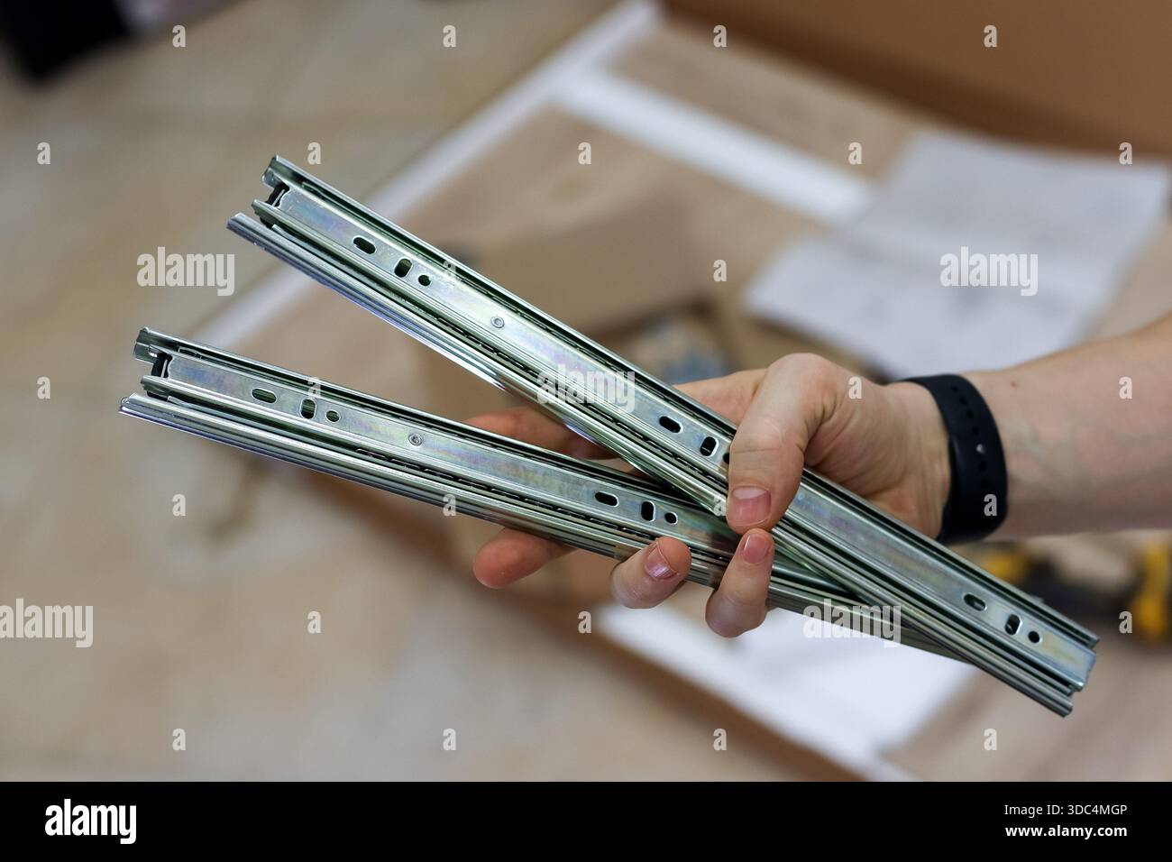 Gros plan de la boulonnerie de glissière de tiroir tenant à la main. Main présentant paire de glissières de tiroir télescopiques métalliques sur fond flou de meubles par Banque D'Images