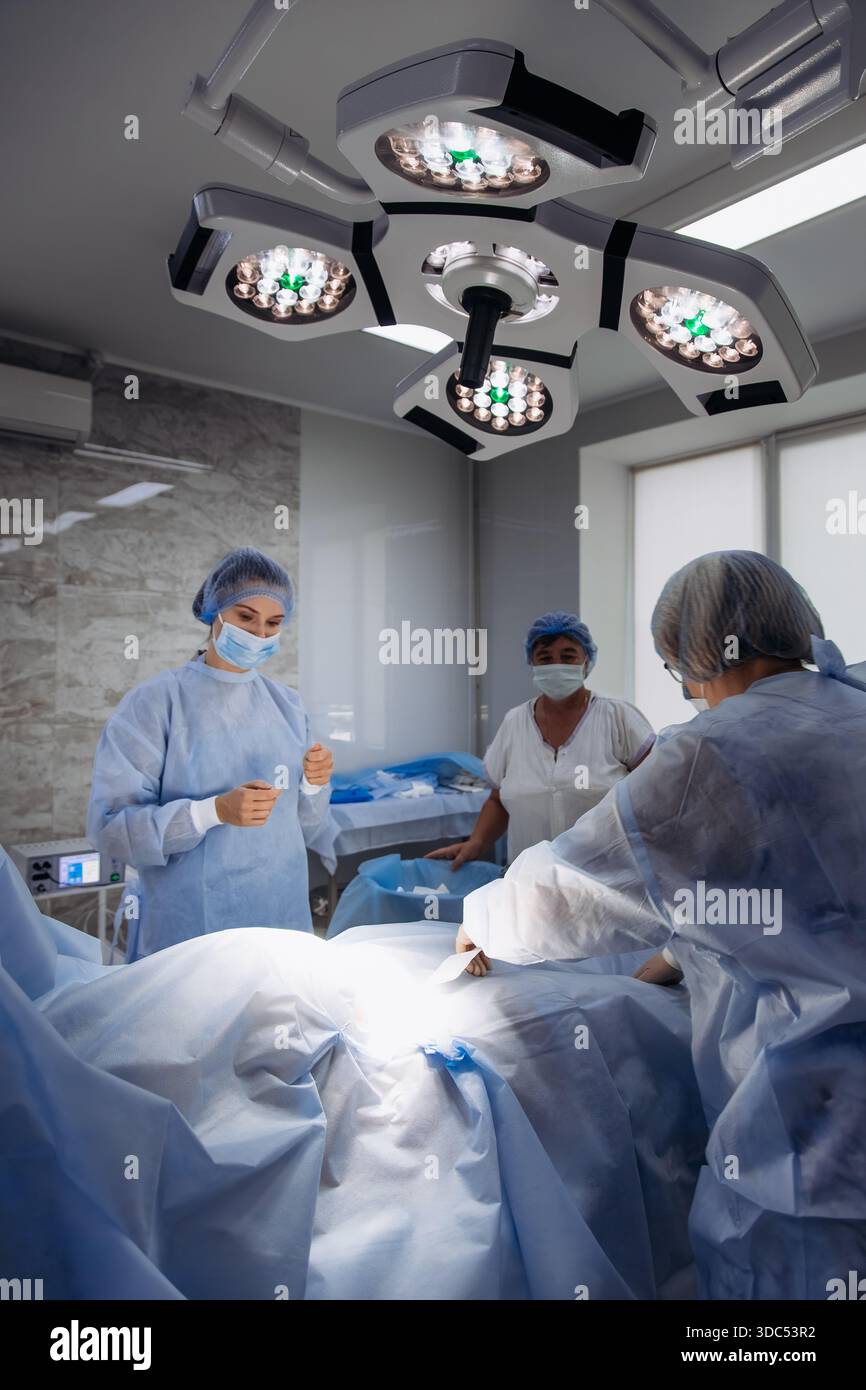 L'équipe chirurgicale dans les gommages et les masques est engagée dans une procédure sous des lumières vives, soulignant la collaboration et la concentration dans un set de salle d'opération stérile Banque D'Images