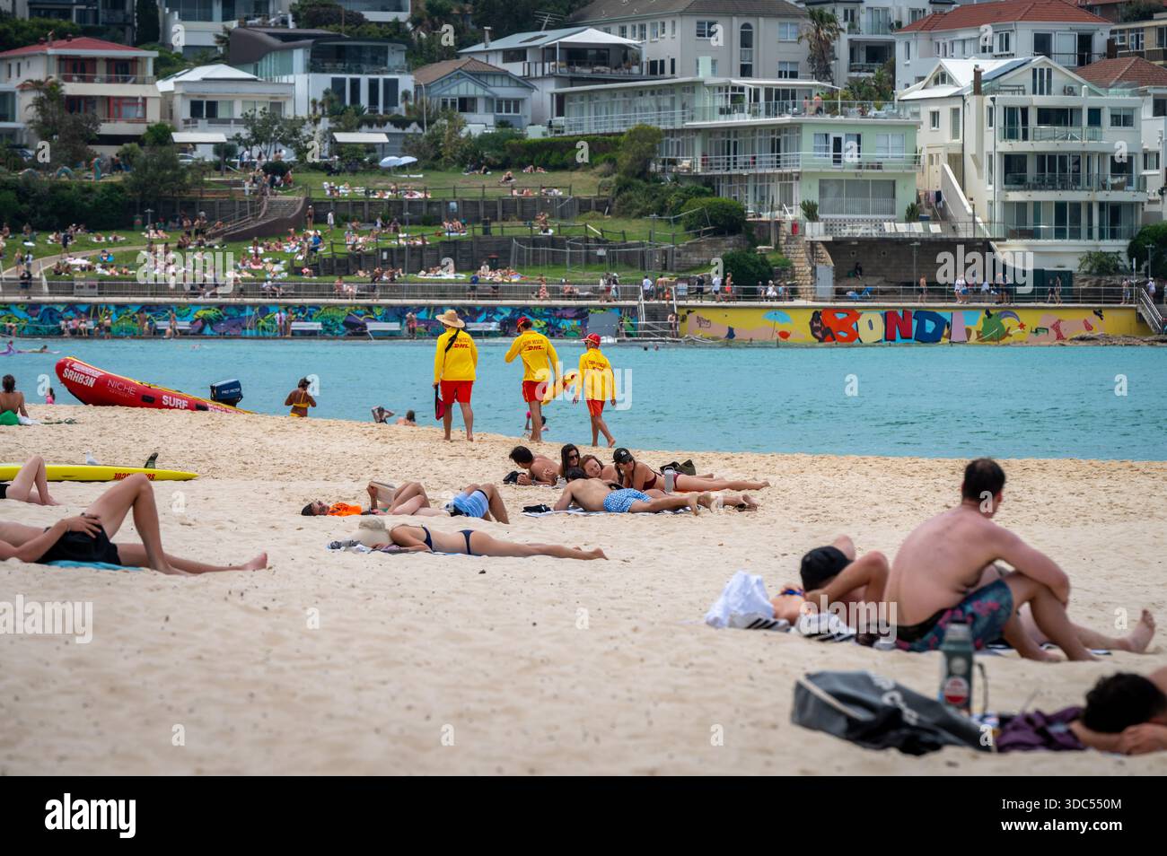 Trois sauveteurs de surf marchent le long du rivage de Bondi Beach pendant un après-midi ensoleillé. Les visiteurs se détendent sur le sable, w / murale murale 'Bondi' est derrière. Banque D'Images