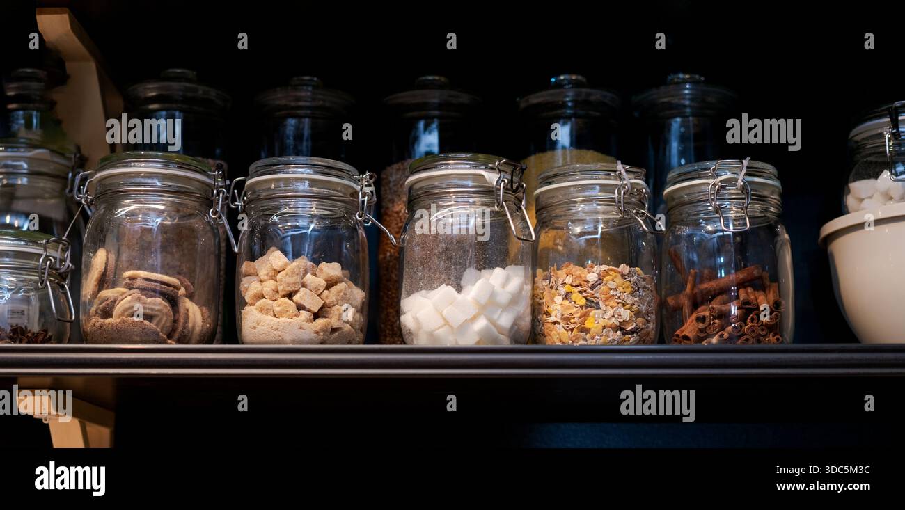 Une étagère pleine de bocaux en verre avec divers aliments à l'intérieur. Les bocaux sont disposés en rangée, certains contenant du sucre, d'autres contenant des épices, et un Banque D'Images