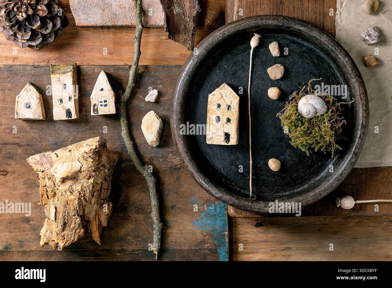 Composition rustique de conception créative plate avec des maisons miniatures en céramique, des galets, coquille, mousse, pomme de pin, plume, brindilles sur fond en bois. Natur Banque D'Images