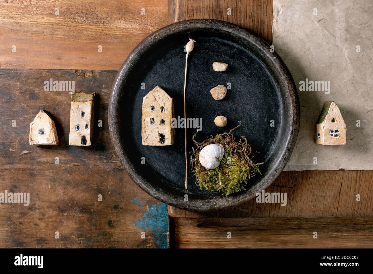 Gros plan bol en céramique noire avec maison en céramique miniature, petits cailloux, nid de mousse avec coquille sur fond de bois rustique. Matériaux naturels artistiques Banque D'Images
