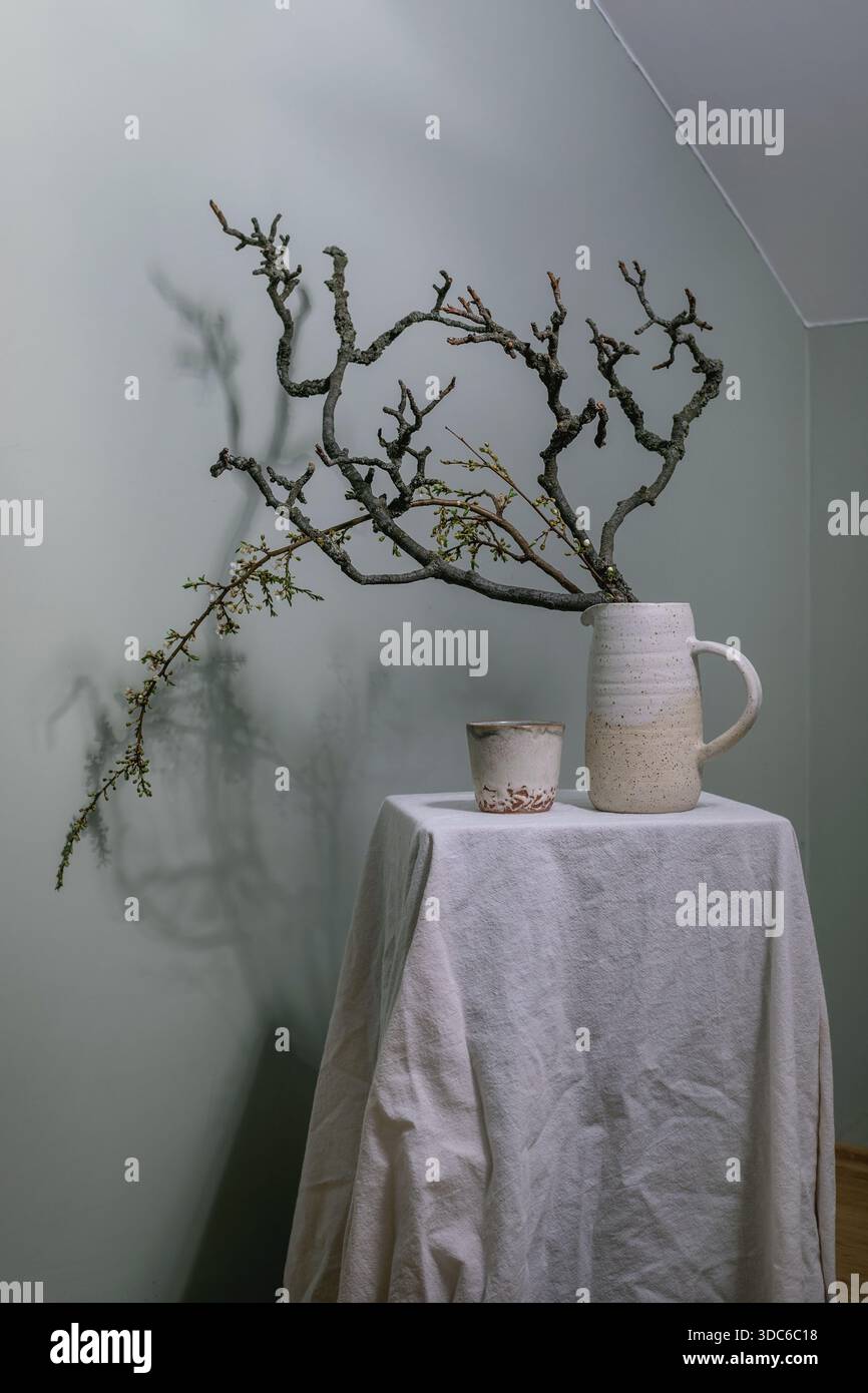 Nature morte minimaliste avec des branches séchées disposées dans un pichet en céramique artisanal, à côté d'une petite tasse sur une table recouverte de tissu blanc contre W vert clair Banque D'Images