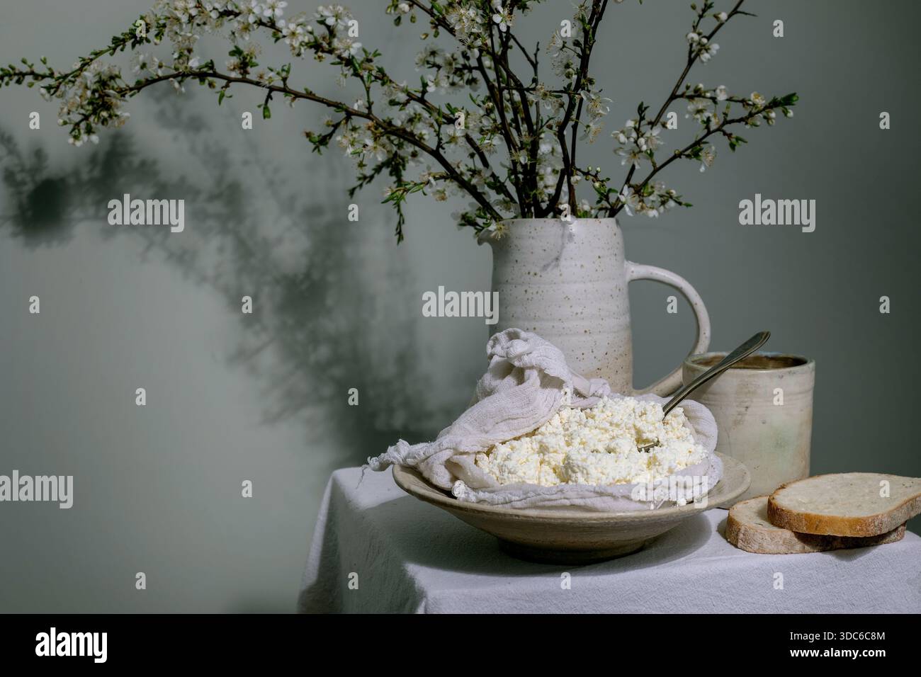 Bol de fromage cottage frais sur un chiffon blanc avec pichet en céramique à pain avec fleurs de printemps contre le mur de menthe. Composition du petit déjeuner de style rustique. Banque D'Images