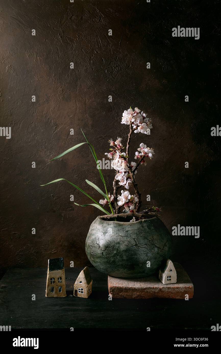 Délicats fleurs de cerisier roses ikebana disposés dans un pot en céramique vert vintage avec des maisons en céramique miniatures sur fond texturé foncé. Moody STIL Banque D'Images