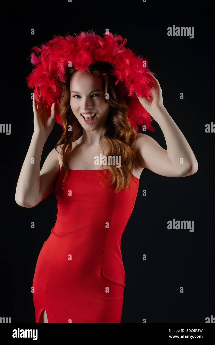 Une femme se tient dans un studio, souriant et posant avec une énergie ludique. Elle porte une robe rouge frappante et un grand chapeau à plumes, exprimant la joie et la confiance dans son expression et sa position. || autorisation du modèle Banque D'Images