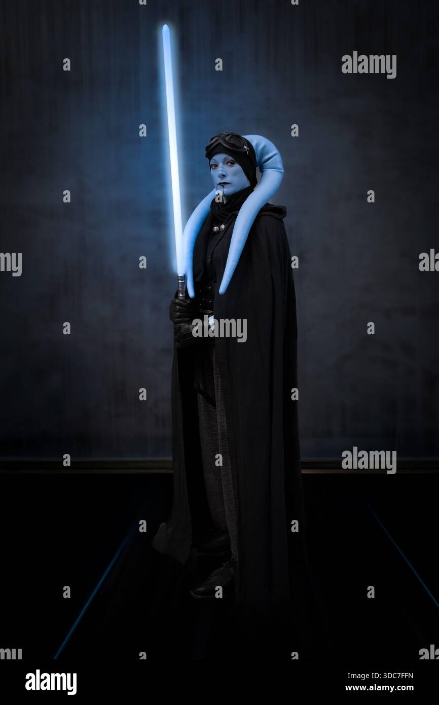 Portrait en pied d'une fille cosplayer habillée en maître Jedi Star Wars Aayla Secura avec un sabre laser bleu et un fond noir Banque D'Images