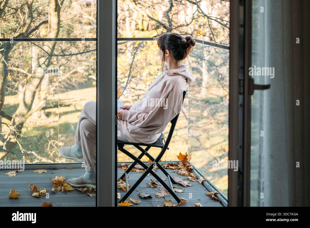 Femme assise sur la chaise sur le balcon lisant livre entouré de feuilles d'automne tombées. Vie lente, solitude urbaine, moments conscients, plaisirs simples. Banque D'Images