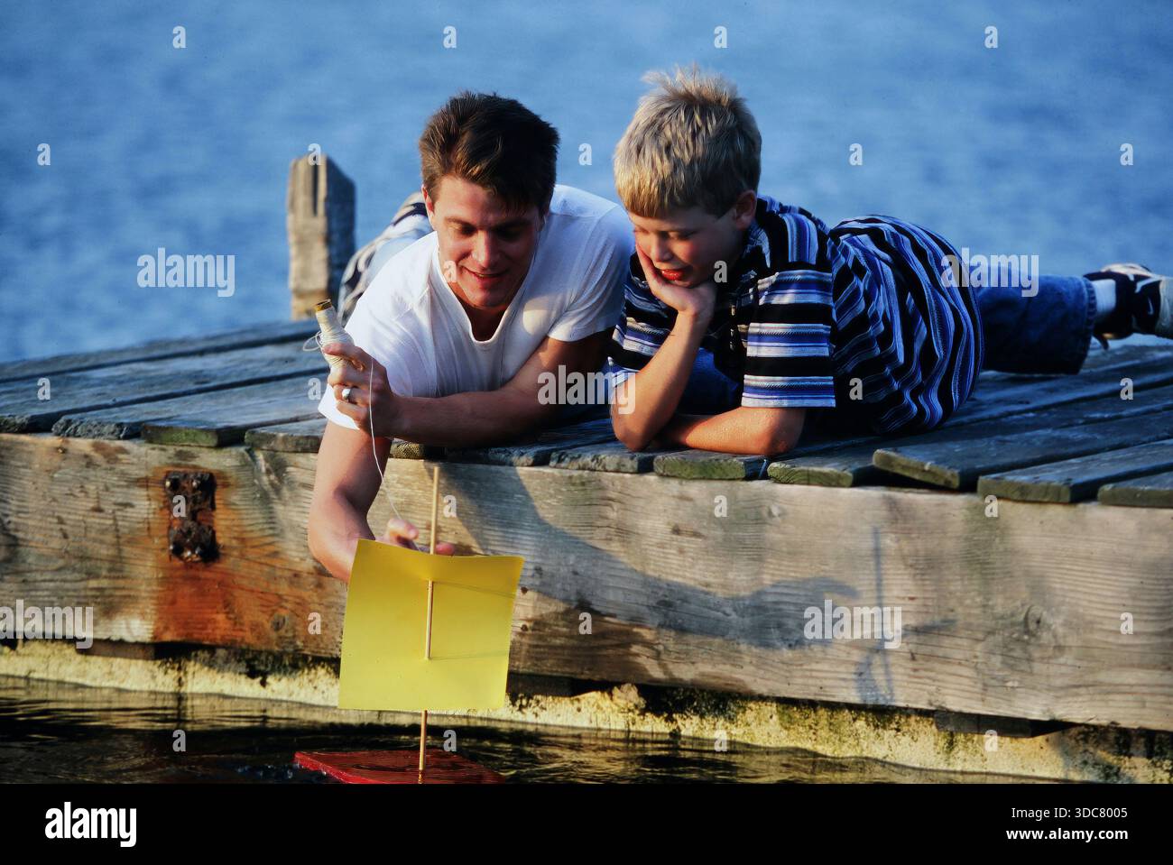 Un père et son fils sont sur un quai près de l'eau, s'amusant comme ils naviguent sur un petit voilier fait maison. Le garçon regarde de près, montrant de l'intérêt pour th Banque D'Images