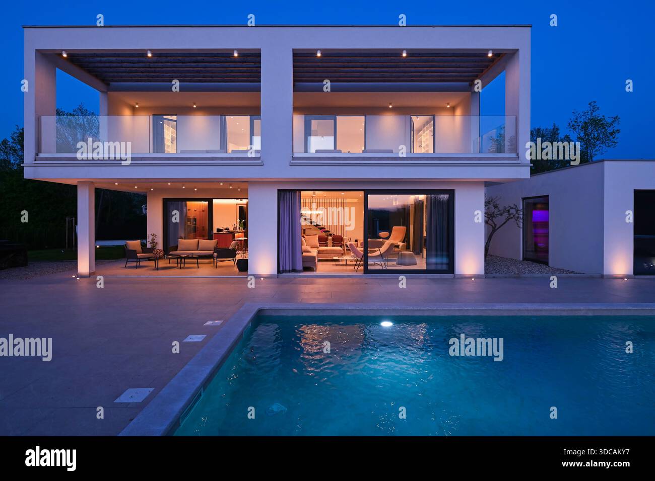 Vue extérieure d'une maison de luxe moderne avec une piscine au crépuscule. Villa blanche illuminée de deux étages avec de grandes fenêtres en verre, un patio, une terrasse et une eau bleue éclatante. Banque D'Images