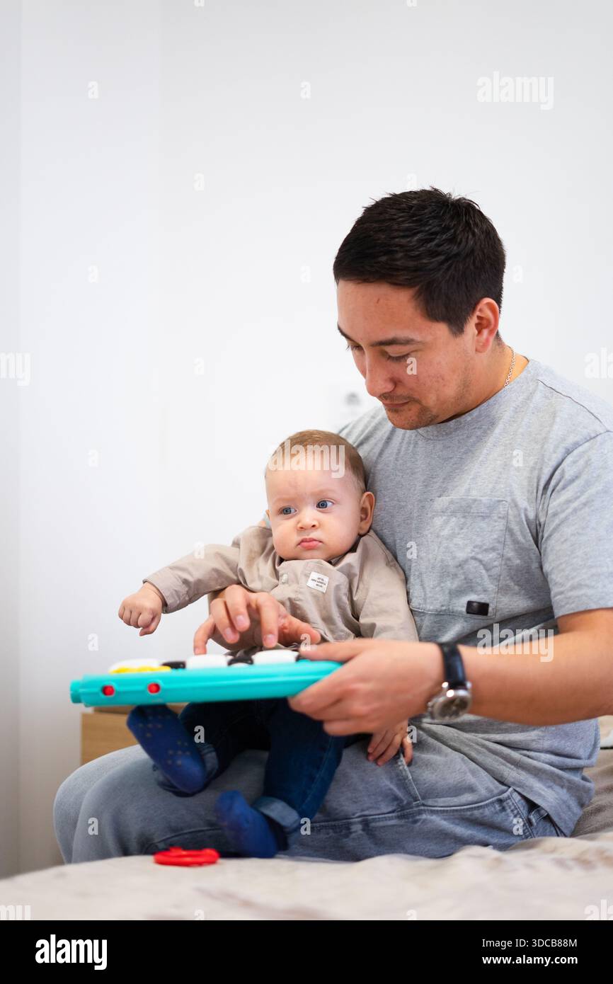 Père latin tenant son bébé fils sur ses genoux, l'aidant à jouer avec un jouet musical coloré, favorisant le développement de la petite enfance Banque D'Images