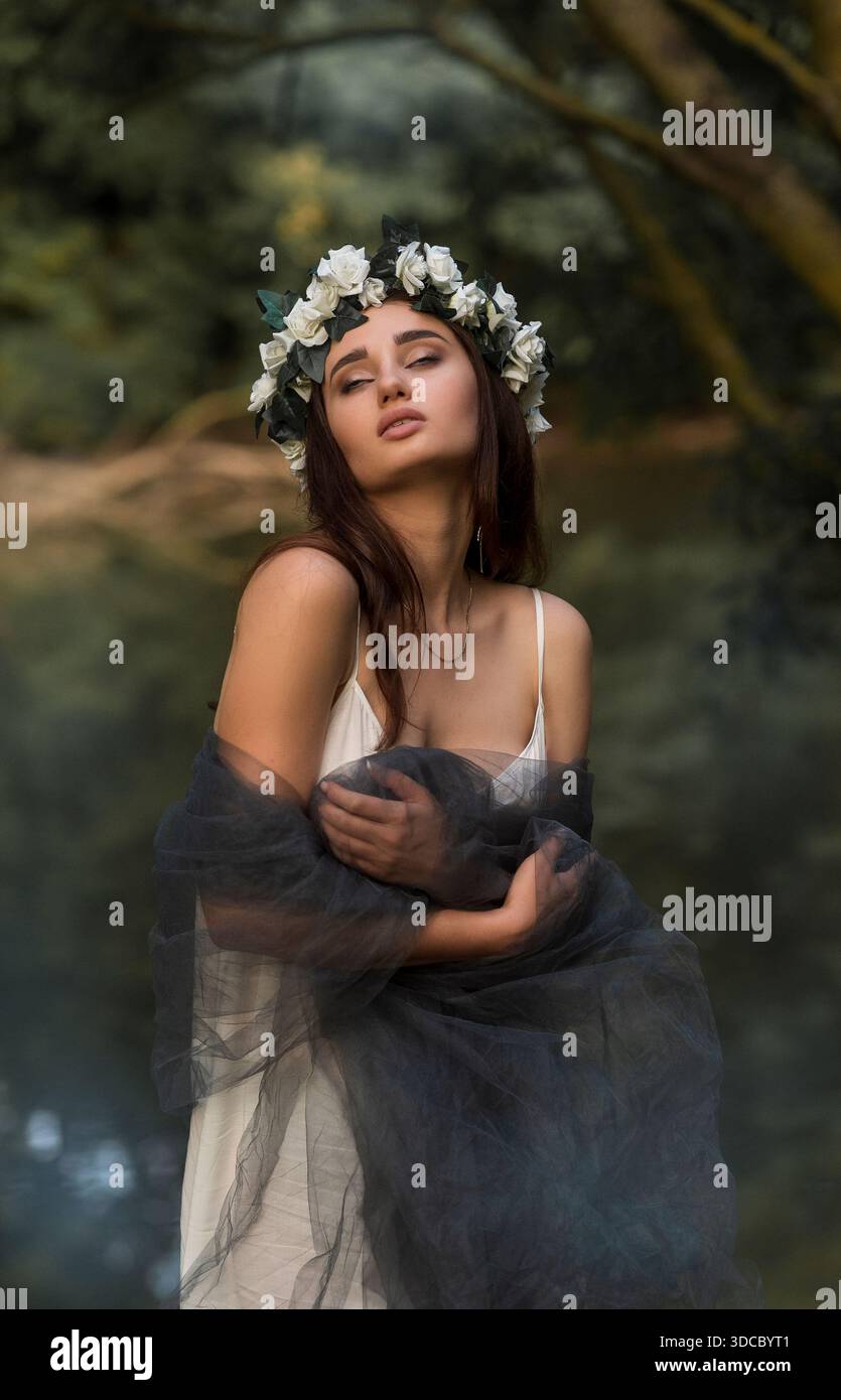 Nymphe forestière dans une robe blanche et couronne de fleurs par un réservoir entouré d'arbres verts. Concept d'art fantastique, beauté de la nature mystique, et cinématographique Banque D'Images