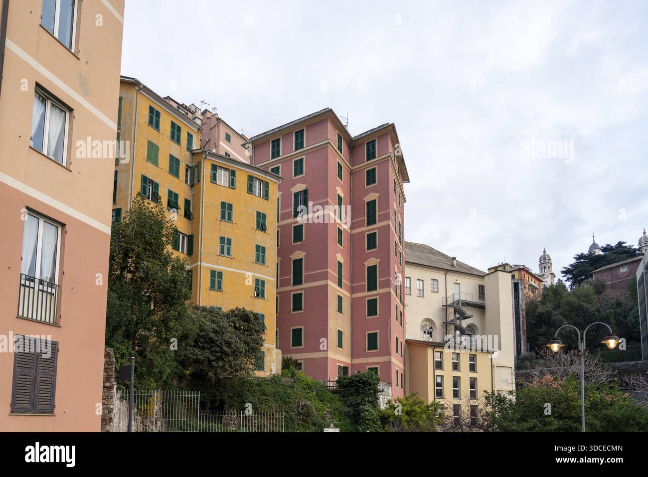 Vue des bâtiments vibrants dans les tons pêche, jaune et rose se dressent sur un fond de verdure luxuriante et un ciel nuageux, Gênes, Ligurie, Italie. Banque D'Images